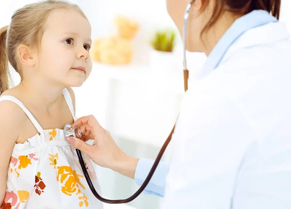 Arzt untersucht ein Kind mit Stethoskop in sonniger Klinik. Glücklich lächelnde Patientin im hellen Kleid ist bei der üblichen ärztlichen Untersuchung — Stockfoto