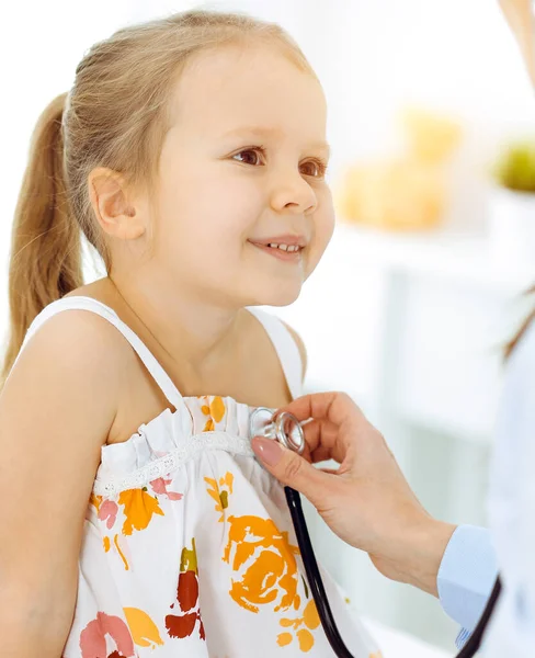 Médecin examinant un enfant par stéthoscope dans une clinique ensoleillée. Heureuse fille souriante patient habillé en robe de couleur vive est à l'inspection médicale habituelle — Photo