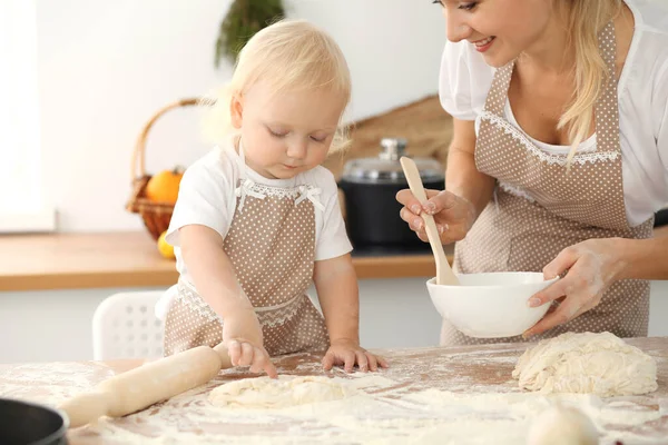 Little girl and her blonde mom in beige aprons playing and laughing while kneading the dough in kitchen. Homemade pastry for bread, pizza or bake cookies. Family fun and cooking concept — Stock Photo, Image