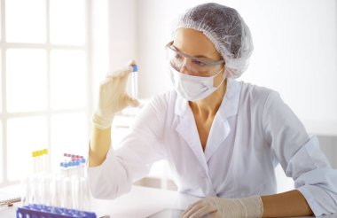 Koruyucu gözlüklü profesyonel kadın bilim adamı güneşli laboratuvarda reaktörlerle araştırma tüpü kullanıyor. Tıp ve araştırma.