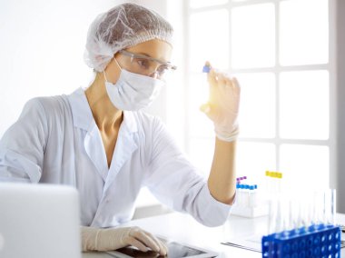 Koruyucu gözlüklü profesyonel kadın bilim adamı güneşli laboratuvarda reaktörlerle araştırma tüpü kullanıyor. Tıp ve araştırma.