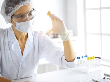 Koruyucu gözlüklü profesyonel kadın bilim adamı güneşli laboratuvarda reaktörlerle araştırma tüpü kullanıyor. Tıp ve araştırma.