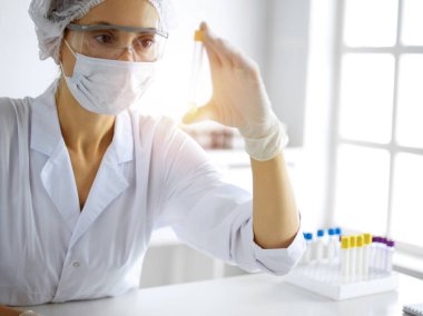 Koruyucu gözlüklü profesyonel kadın bilim adamı güneşli laboratuvarda reaktörlerle araştırma tüpü kullanıyor. Tıp ve araştırma.