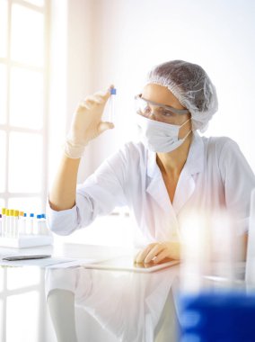 Koruyucu gözlüklü profesyonel kadın bilim adamı güneşli laboratuvarda reaktörlerle araştırma tüpü kullanıyor. Tıp ve araştırma.