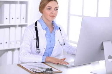 Hastanedeki genç bayan doktor masaüstü bilgisayar monitörüne bakıyor. Doktor ilaç geçmişi ve sınav sonuçlarını kontrol ediyor. Tıp ve sağlık hizmetleri kavramı