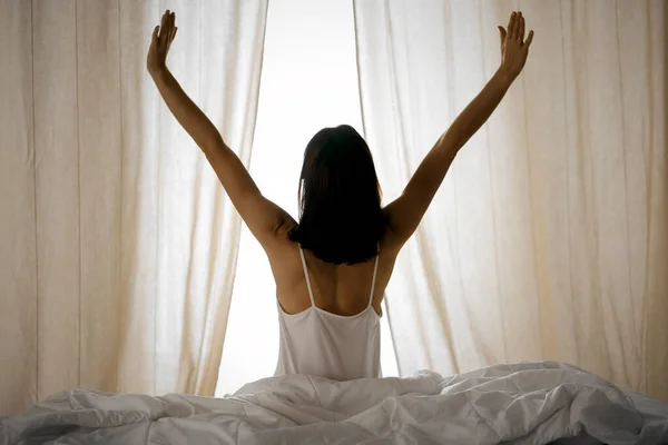 Woman stretching hands in bed after wake up, sun flare . Brunette entering a day happy and relaxed after good night sleep and back view. Concept of a new day and joyful weekend