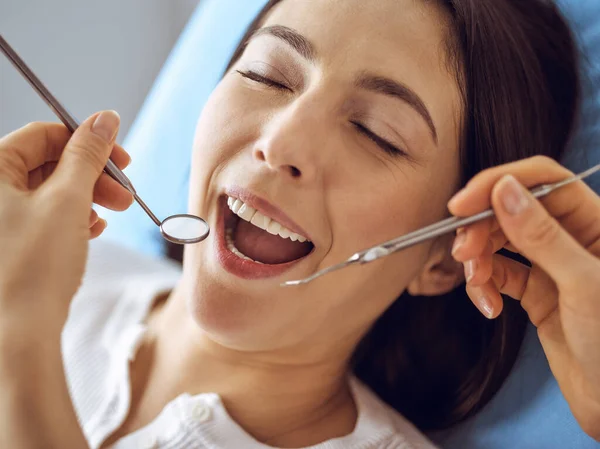 Uśmiechnięta brunetka badana przez dentystę w klinice dentystycznej. Ręce lekarza trzymającego instrumenty stomatologiczne w pobliżu ust pacjenta. Zdrowe zęby i koncepcja medycyny — Zdjęcie stockowe