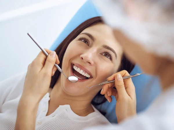 Lachende brunette vrouw wordt onderzocht door tandarts in de tandheelkundige kliniek. Handen van een arts met tandheelkundige instrumenten in de buurt van de mond van patiënten. Gezonde tanden en geneeskundeconcept — Stockfoto