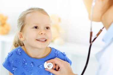 Güneşli klinikte bir çocuğu steteskopla muayene eden bir doktor. Mavi elbiseli mutlu gülümseyen kız hasta her zamanki sağlık kontrolünde.