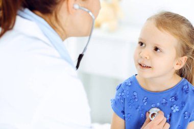Güneşli klinikte bir çocuğu steteskopla muayene eden bir doktor. Mavi elbiseli mutlu gülümseyen kız hasta her zamanki sağlık kontrolünde.