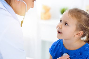 Güneşli klinikte bir çocuğu steteskopla muayene eden bir doktor. Mavi elbiseli mutlu gülümseyen kız hasta her zamanki sağlık kontrolünde.