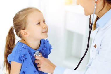 Güneşli klinikte bir çocuğu steteskopla muayene eden bir doktor. Mavi elbiseli mutlu gülümseyen kız hasta her zamanki sağlık kontrolünde.