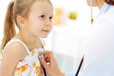 Güneşli klinikte bir çocuğu steteskopla muayene eden bir doktor. Parlak renkli elbise giymiş mutlu gülümseyen kız hasta her zamanki sağlık kontrolünde.