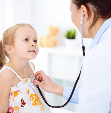 Güneşli klinikte bir çocuğu steteskopla muayene eden bir doktor. Parlak renkli elbise giymiş mutlu gülümseyen kız hasta her zamanki sağlık kontrolünde.