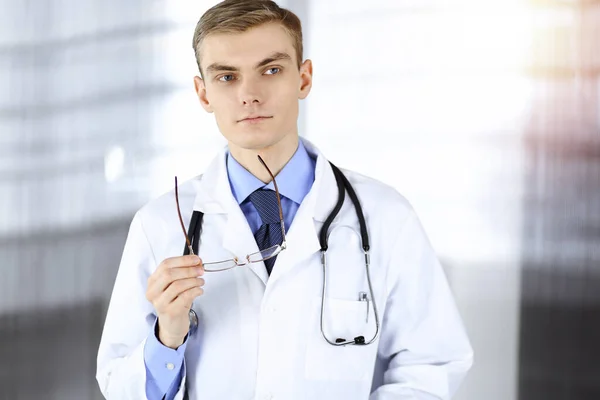 El doctor, con gafas y estetoscopio, está de pie en su soleado consultorio del hospital. Servicio médico perfecto en la clínica. Futuro feliz en medicina y salud —  Fotos de Stock