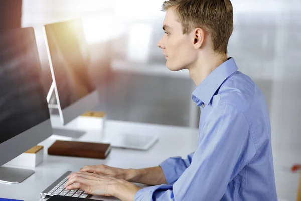 Jeune homme d'affaires prospère travaille avec l'ordinateur, assis au bureau dans un bureau moderne ensoleillé. Prise de vue de l'entrepreneur masculin ou it-spécialiste sur le lieu de travail. Concept d'entreprise — Photo