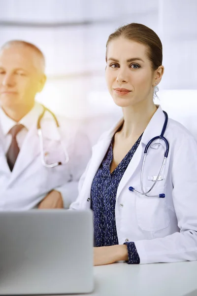 Elderly doctor and his female colleague discussing current disease therapy while sitting at working place in sunny clinic. Teamwork in medicine