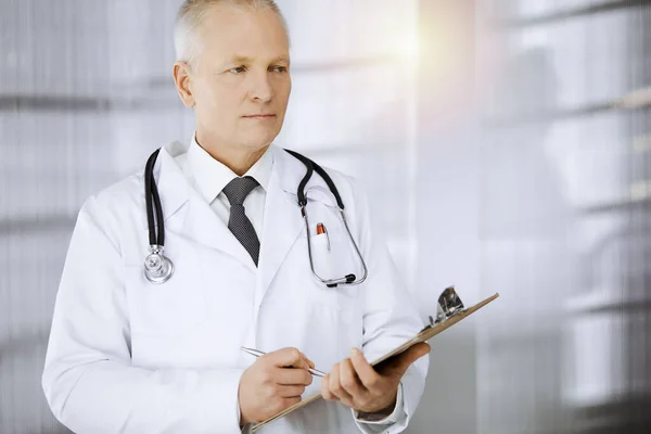 나이든 자신감 있는 의사가 똑바로 서서 맑은 병원에서 클립보드를 사용하고 있습니다. 완벽 한 의료 서비스죠. 의학적 개념 — 스톡 사진