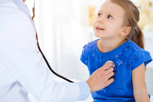 Врач осматривает ребенка стетоскопом в солнечной клинике. Счастливая улыбающаяся пациентка в синем платье при обычном медицинском осмотре — стоковое фото