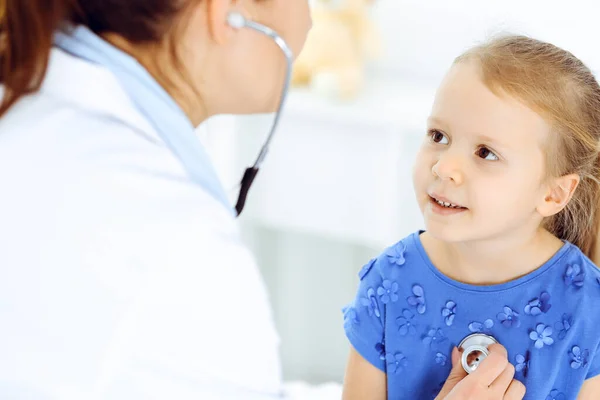 Doutor examinando uma criança por estetoscópio na clínica ensolarada. Feliz sorrindo paciente menina vestida de vestido azul é na inspeção médica habitual — Fotografia de Stock