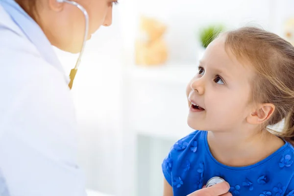 Doutor examinando uma criança por estetoscópio na clínica ensolarada. Feliz sorrindo paciente menina vestida de vestido azul é na inspeção médica habitual — Fotografia de Stock