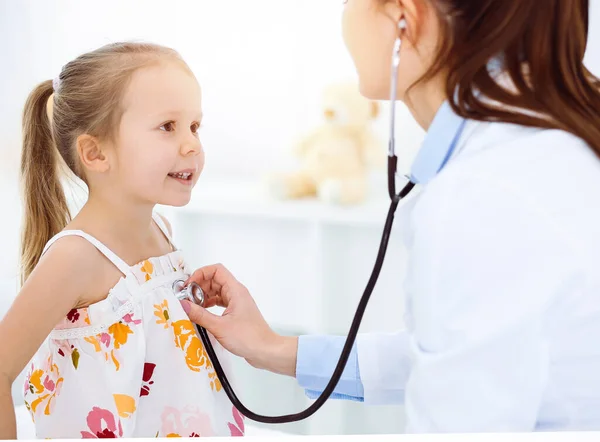 Arzt untersucht ein Kind mit Stethoskop in sonniger Klinik. Glücklich lächelnde Patientin im hellen Kleid ist bei der üblichen ärztlichen Untersuchung — Stockfoto