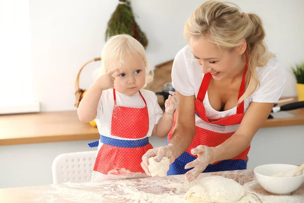 Klein meisje en haar blonde moeder in rode schorten spelen en lachen terwijl ze het deeg kneden in de keuken. Zelfgebakken gebak voor brood, pizza of koekjes bakken. Familie plezier en koken concept — Stockfoto