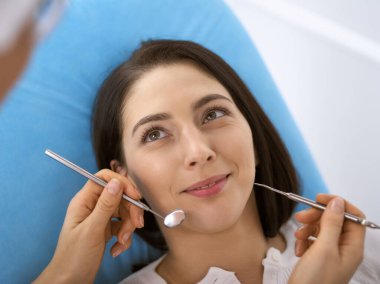 Gülümseyen esmer kadın diş hekimi tarafından muayene ediliyor. Hastanın ağzının yanında diş aletleri tutan bir doktorun elleri. Sağlıklı diş ve ilaç konsepti