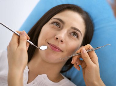 Gülümseyen esmer kadın diş hekimi tarafından muayene ediliyor. Hastanın ağzının yanında diş aletleri tutan bir doktorun elleri. Sağlıklı diş ve ilaç konsepti