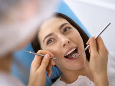 Gülümseyen esmer kadın diş hekimi tarafından muayene ediliyor. Hastanın ağzının yanında diş aletleri tutan bir doktorun elleri. Sağlıklı diş ve ilaç konsepti