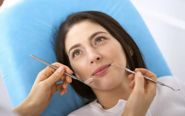 Lachende brunette vrouw wordt onderzocht door tandarts in de tandheelkundige kliniek. Handen van een arts met tandheelkundige instrumenten in de buurt van de mond van patiënten. Gezonde tanden en geneeskundeconcept — Stockfoto