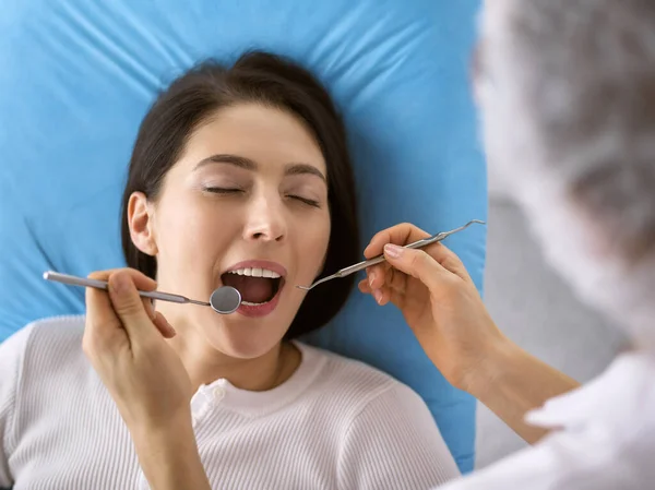 Leende brunett kvinna som undersöks av tandläkare på tandkliniken. Händerna på en läkare som håller dentala instrument nära patientens mun. Friska tänder och medicin koncept — Stockfoto