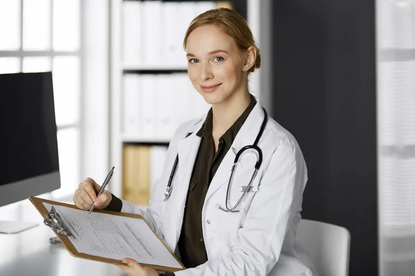 Alegre sonriente doctora usando portapapeles en la clínica. Retrato de mujer médica amigable en el lugar de trabajo. Servicio médico perfecto en el hospital. Concepto de medicina — Foto de Stock