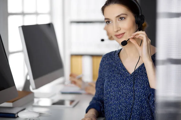 Donna d'affari latinoamericana che parla in cuffia mentre è in carica. Call center e diversi gruppi di persone nel mondo degli affari — Foto Stock