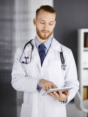 Dost canlısı kızıl sakallı doktor çalıştığı yerdeki klinikte tablet bilgisayar kullanıyor. Hastane ve tıp konseptinde mükemmel bir tıbbi hizmet.