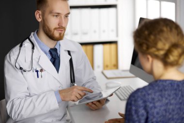 Dost canlısı kızıl sakallı doktor ve hasta kadın klinikte oturup tablet bilgisayar kullanırken sağlık muayenesini tartışıyorlar. Tıp konsepti