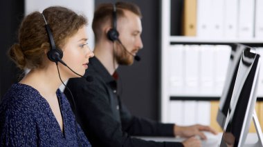 Modern ofiste kızıl sakallı bir meslektaşla otururken iş kadını kulaklıkla konuşuyor. Tele pazarlama ve müşteri hizmetleri. Çağrı merkezinde çeşitli insanlar grubu