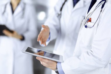 Bilinmeyen bir grup doktor hastane ofisinde dururken bazı tıbbi isim kayıtlarını kontrol etmek için bir bilgisayar tableti kullanıyor. Doktorlar hastaları muayene etmeye ve yardım etmeye hazır. Tıbbi yardım, sigorta