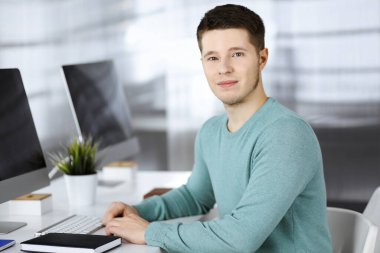 Yeşil kazak giyen kendine güvenen genç işadamı, yeni projesi üzerinde çalışırken modern bir ofisin masasında oturuyor. Erkek girişimcinin ya da iş adamının vesikalık fotoğrafı