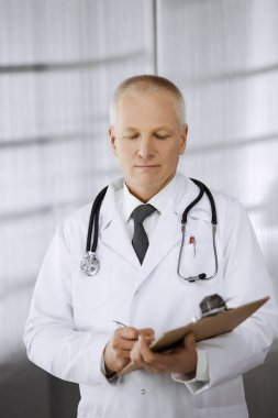 Yaşlı, kendine güvenen bir doktor ayakta duruyor ve klinikte pano kullanıyor. Mükemmel bir tıbbi servis. Tıp ve sağlık hizmetleri kavramı