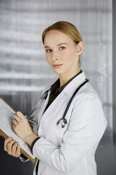 Friendly smiling female doctor using clipboard in clinic. Portrait of friendly physician woman at work. Medical service in hospital. Medicine concept