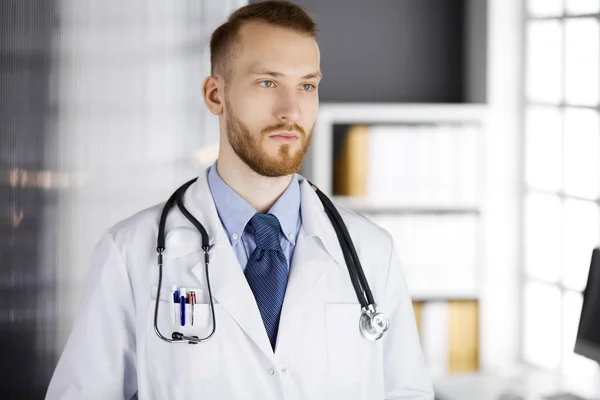Рыжебородатый врач, стоящий прямо в клинике рядом с рабочим местом. Портрет врача. Медицина и здравоохранение — стоковое фото