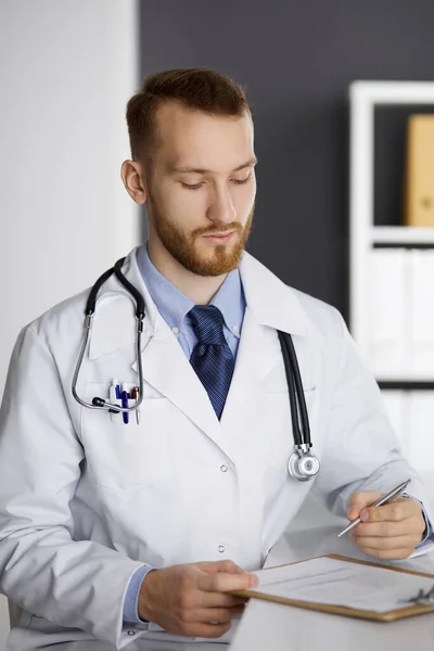 Φιλικός γιατρός με κόκκινα γένια κάθεται και γράφει στο πρόχειρο στην κλινική. Τέλεια ιατρική υπηρεσία στο νοσοκομείο. Έννοια ιατρικής — Φωτογραφία Αρχείου