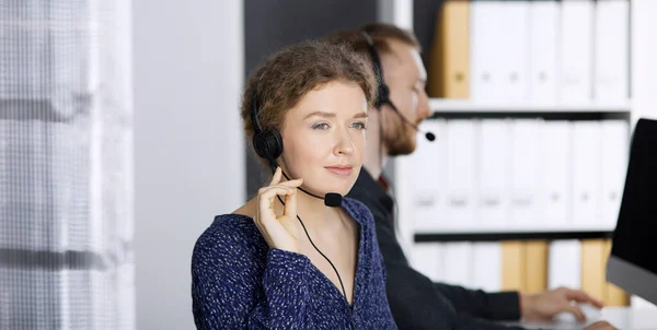 Femme d'affaires parlant par casque tout en étant assis avec un collègue à barbe rouge dans le bureau moderne. Télémarketing et service à la clientèle. Groupe de personnes diversifiées dans le centre d'appels — Photo