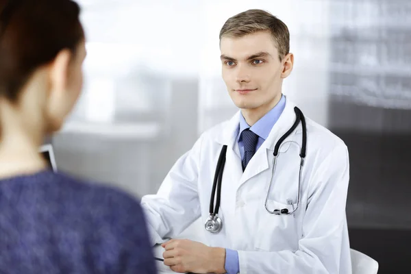 Dokter man, met een stethoscoop, luistert naar zijn patiënt, terwijl hij aan het bureau in het ziekenhuis zit. Perfecte medische dienst in de kliniek. Gelukkige toekomst in de geneeskunde en de gezondheidszorg — Stockfoto