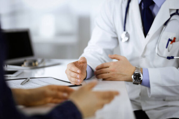 A doctor is talking to his patient, while sitting together at the desk in the cabinet in a hospital. Physician using clipboard for filling up medication history records. Perfect medical service in