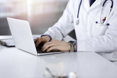 Bilinmeyen bir erkek doktor karanlık bir klinikte dizüstü bilgisayarla oturuyor ve çalışıyor. Arka planda ışık parıltısı var.