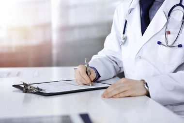 Bilinmeyen bir erkek doktor, karanlık bir klinikte tıbbi geçmişi olan bir panoyla oturuyor ve çalışıyor. Arka planda ışık parıltısı var.