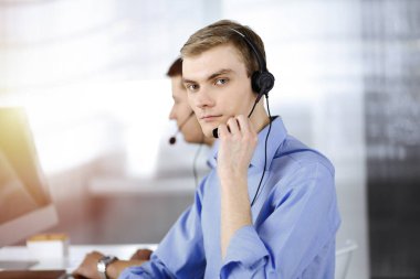 Kulaklıklı iki genç adam güneşli, modern bir ofiste masada oturup müşterileri dinliyor. İş yerindeki çağrı merkezi operatörleri