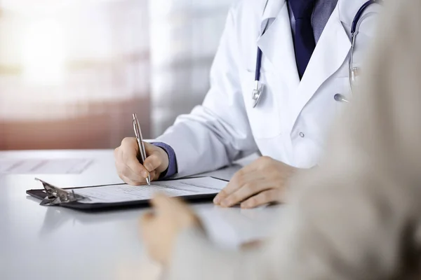 Médico masculino desconocido y mujer paciente discutiendo algo mientras está sentado en una clínica oscura y usando portapapeles, resplandor de luz en el fondo — Foto de Stock